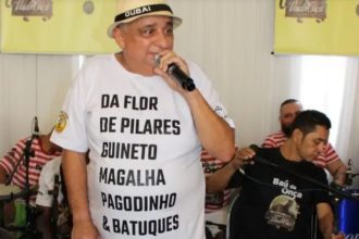 Paulo Onça, sambista, em um evento com um microfone na mão, usando uma camiseta branca com a frase "Da Flor de Pilares Guineto Magalha Pagodinho & Batuques".