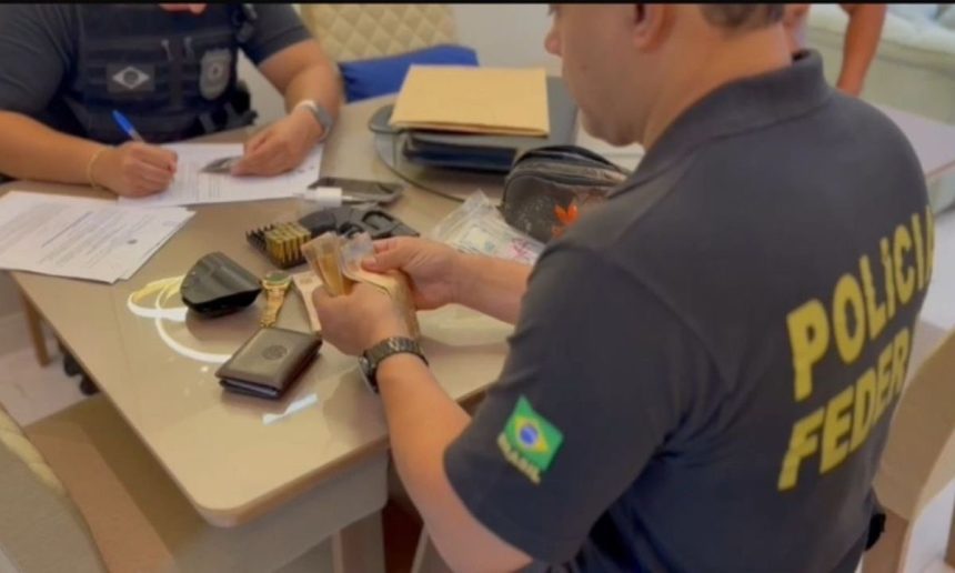 Policial da PF inspeciona dinheiro apreendido durante operação em uma mesa com outros objetos.