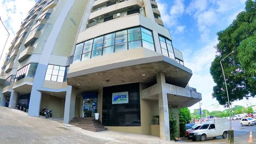 Prédio da CDL Manaus com fachada moderna em tons de cinza e detalhes em azul, localizado em uma rua com árvores e outros edifícios ao fundo.