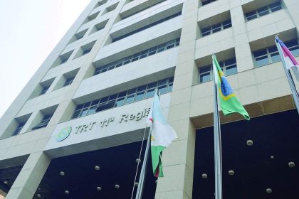 Prédio da Justiça do Trabalho com a bandeira do Brasil e outras bandeiras, em frente a um céu azul claro.
