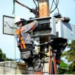 Técnicos trabalhando em um poste de energia com equipamentos de segurança, realizando manutenção na rede elétrica.
