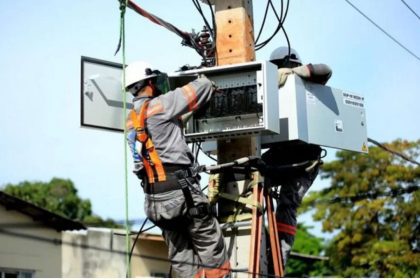 Técnicos trabalhando em um poste de energia com equipamentos de segurança, realizando manutenção na rede elétrica.