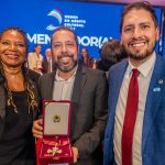 Três pessoas em um evento de premiação, sendo duas mulheres e um homem. O homem está segurando uma medalha em uma caixa vermelha.