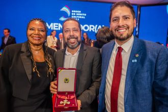 Três pessoas em um evento de premiação, sendo duas mulheres e um homem. O homem está segurando uma medalha em uma caixa vermelha.