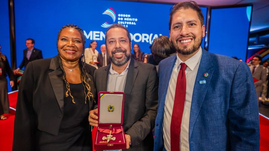 Três pessoas em um evento de premiação, sendo duas mulheres e um homem. O homem está segurando uma medalha em uma caixa vermelha.