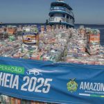 Um barco cheio de pacotes de alimentos, sinalizado com 'Operação Cheia 2025' e 'Defesa Civil Amazonas', para ajudar vítimas de enchentes no Amazonas.