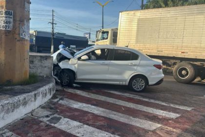 Um carro branco, aparentemente acidentado, encontra-se em um cruzamento, com a frente amassada e sinais de colisão.