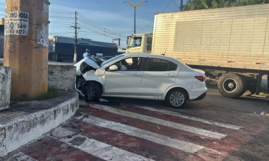 Um carro branco, aparentemente acidentado, encontra-se em um cruzamento, com a frente amassada e sinais de colisão.