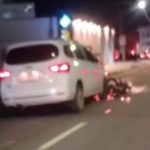 Um carro branco parado em uma rua à noite com uma motocicleta presa embaixo, após um acidente.