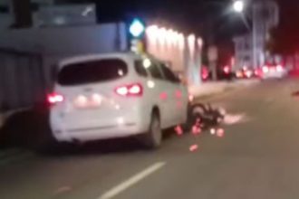 Um carro branco parado em uma rua à noite com uma motocicleta presa embaixo, após um acidente.
