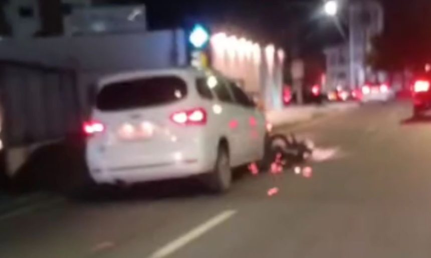 Um carro branco parado em uma rua à noite com uma motocicleta presa embaixo, após um acidente.