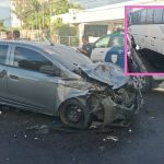 Um carro de passeio cinza com a frente amassada após um acidente com um micro-ônibus branco, que aparece parcialmente na imagem.
