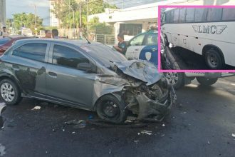 Um carro de passeio cinza com a frente amassada após um acidente com um micro-ônibus branco, que aparece parcialmente na imagem.