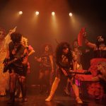 Um grupo de artistas em um palco, com iluminação laranja e um clima festivo, se apresentam durante uma performance teatral.