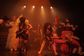 Um grupo de artistas em um palco, com iluminação laranja e um clima festivo, se apresentam durante uma performance teatral.