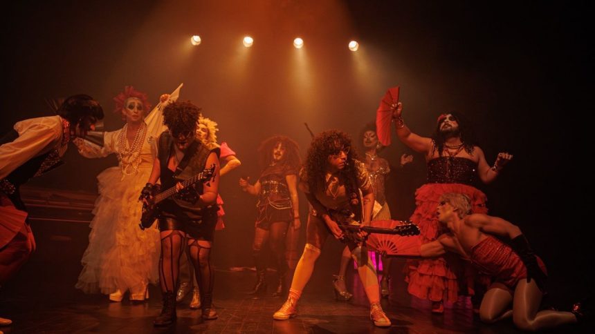 Um grupo de artistas em um palco, com iluminação laranja e um clima festivo, se apresentam durante uma performance teatral.