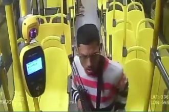 Um homem com roupa listrada branca e vermelha e óculos, aparentemente em um ônibus, visto de frente.