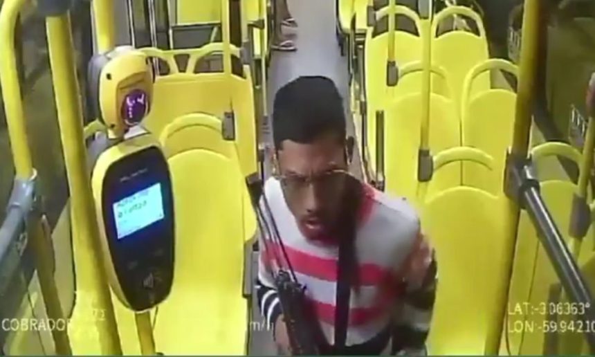 Um homem com roupa listrada branca e vermelha e óculos, aparentemente em um ônibus, visto de frente.