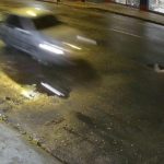 Um homem deitado no chão, em uma rua escura, com um carro passando em alta velocidade. Parece haver um acidente.