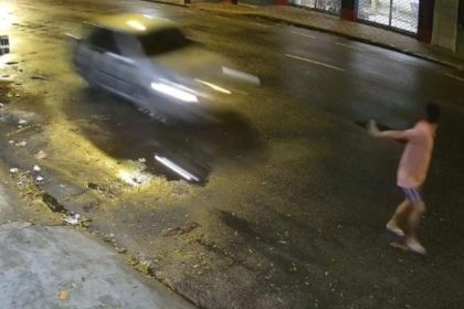 Um homem deitado no chão, em uma rua escura, com um carro passando em alta velocidade. Parece haver um acidente.