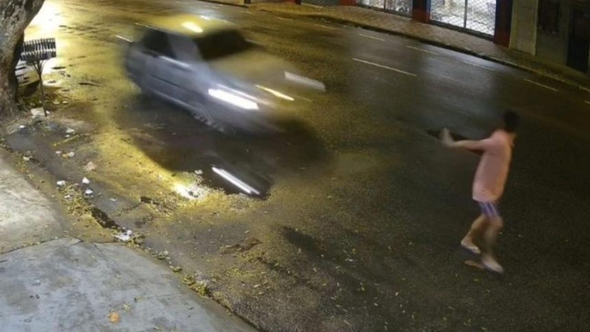 Um homem deitado no chão, em uma rua escura, com um carro passando em alta velocidade. Parece haver um acidente.