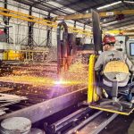 Um trabalhador opera uma máquina de corte a laser em um ambiente industrial, com faíscas voando e um pedaço de metal sendo cortado.