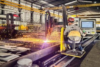 Um trabalhador opera uma máquina de corte a laser em um ambiente industrial, com faíscas voando e um pedaço de metal sendo cortado.