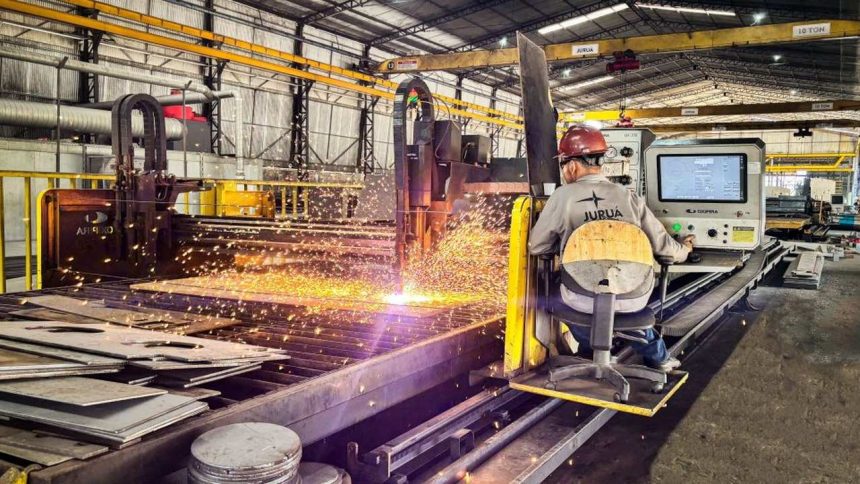 Um trabalhador opera uma máquina de corte a laser em um ambiente industrial, com faíscas voando e um pedaço de metal sendo cortado.