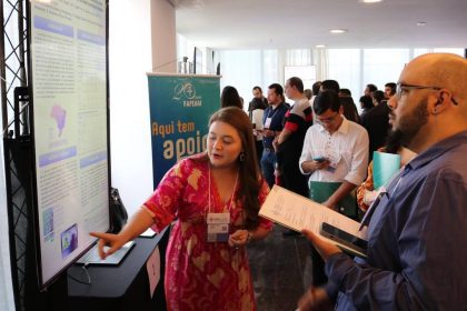 Uma mulher aponta para uma apresentação sobre oncologia em um congresso científico, enquanto um homem a escuta e segura um tablet.