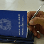 Imagem colorida mostra carteiras de trabalho com uma mão segurando uma caneta