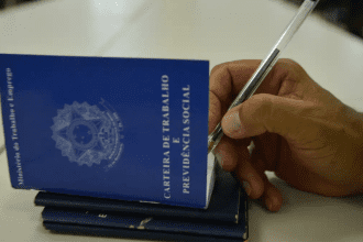 Imagem colorida mostra carteiras de trabalho com uma mão segurando uma caneta