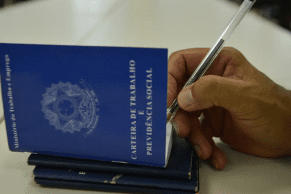 Imagem colorida mostra carteiras de trabalho com uma mão segurando uma caneta