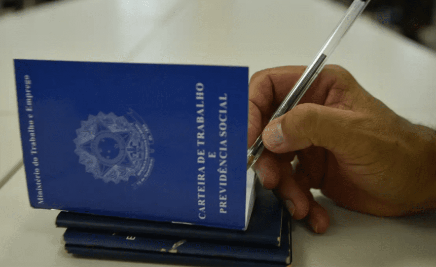 Imagem colorida mostra carteiras de trabalho com uma mão segurando uma caneta