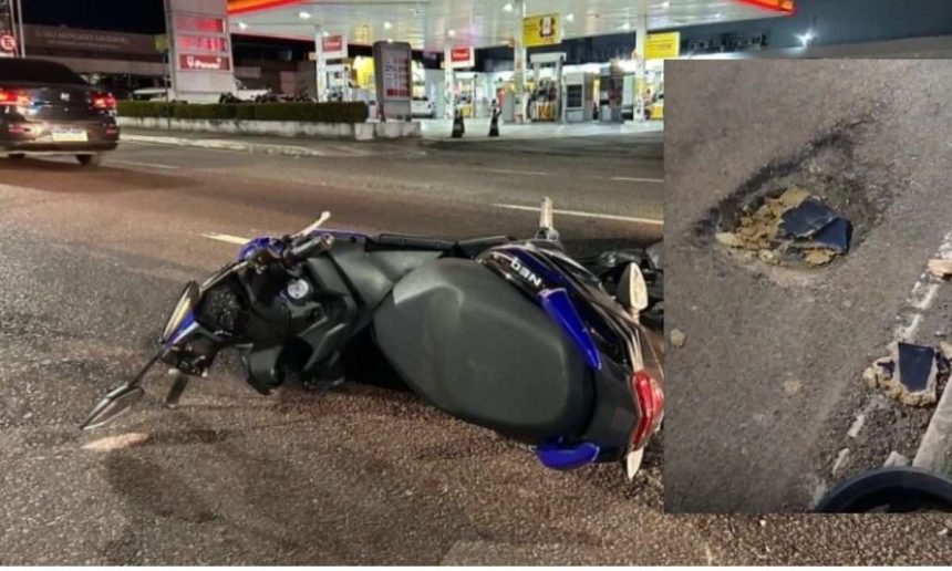 Acidente com moto em avenida de Manaus após buraco, com buraco na pista e posto de gasolina ao fundo.