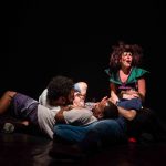 Apresentação teatral com figurinos coloridos e maquiagem de palhaço em palco escuro.