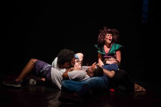 Apresentação teatral com figurinos coloridos e maquiagem de palhaço em palco escuro.