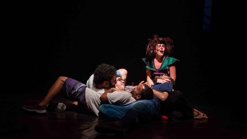 Apresentação teatral com figurinos coloridos e maquiagem de palhaço em palco escuro.