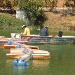 Barcos no lago: imagem de três pessoas em um barco, próximo a estruturas flutuantes e margem.