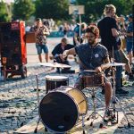 Baterista tocando ao ar livre em evento musical, com público ao fundo.