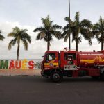 Caminhão do Corpo de Bombeiros em frente a placa 'Eu Amo Maués' com palmeiras ao fundo.