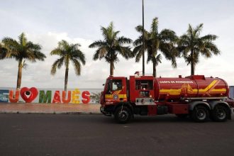 Caminhão do Corpo de Bombeiros em frente a placa 'Eu Amo Maués' com palmeiras ao fundo.
