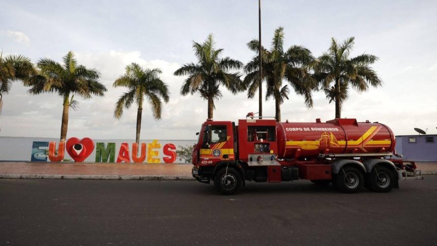 Caminhão do Corpo de Bombeiros em frente a placa 'Eu Amo Maués' com palmeiras ao fundo.