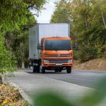 Caminhão laranja Volkswagen Delivery em estrada arborizada.