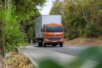 Caminhão laranja Volkswagen Delivery em estrada arborizada.
