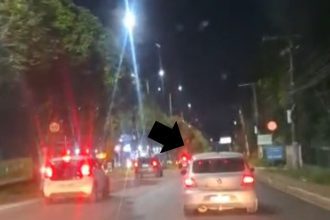 Carros em movimento na Avenida do Turismo à noite, com luzes vermelhas acesas.