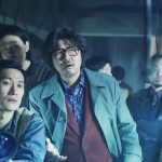 Cena do filme Na Teia da Aranha com elenco em ambiente fechado.