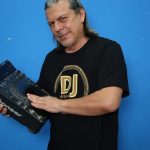 DJ segurando equipamento de som, com camiseta 'DJ Artele' e fundo azul.