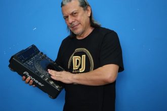 DJ segurando equipamento de som, com camiseta 'DJ Artele' e fundo azul.
