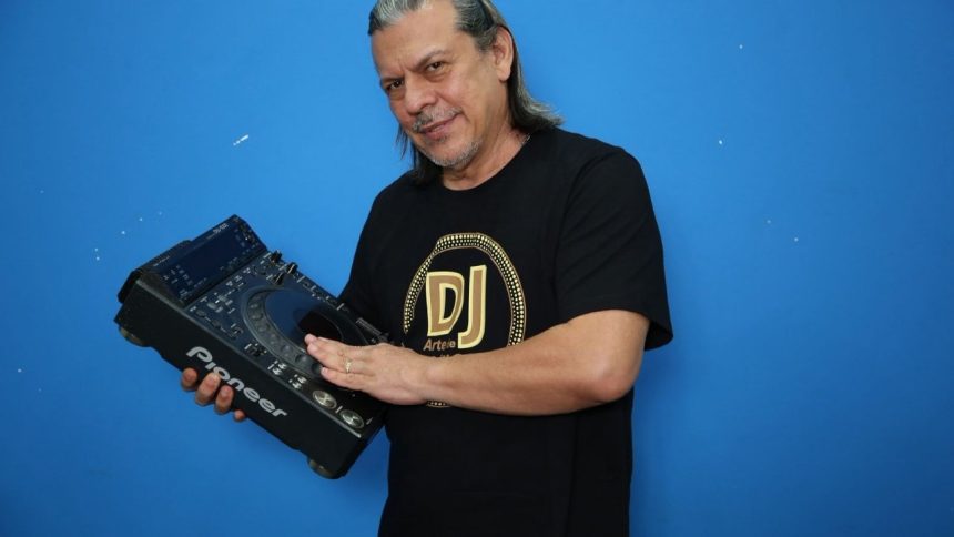 DJ segurando equipamento de som, com camiseta 'DJ Artele' e fundo azul.