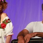 Dois artistas em um palco, uma mulher sorrindo e um homem com expressão relaxada.